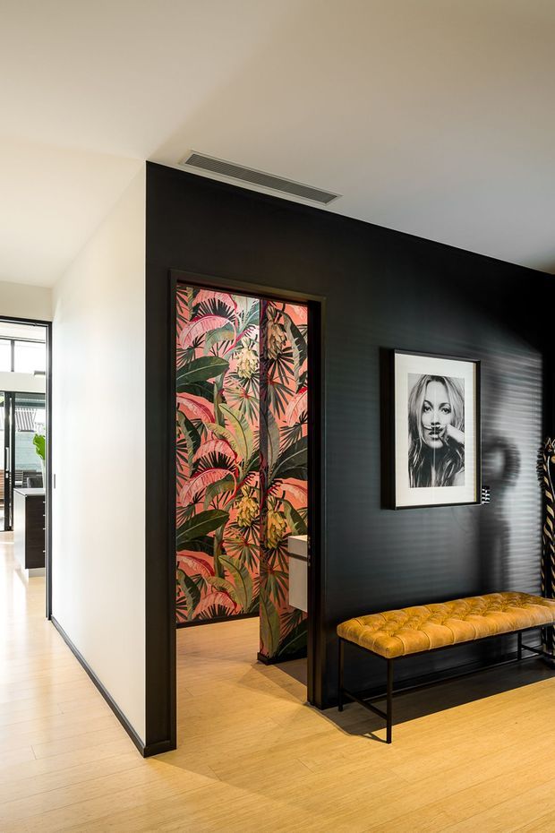 A stylish residential hallway or entry area with a dramatic black feature wall, concealed doorway clad in colourful tropical botanical wallpaper, framed black-and-white artwork, and a mustard-yellow tufted bench on black metal legs. Light timber flooring and white walls create contrast, while a glazed opening at left reveals an adjacent interior space.