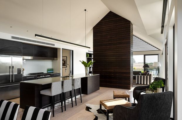 Open-plan contemporary kitchen and living area with a dark timber-clad, gable-shaped feature wall dividing the spaces. The kitchen has dark fixed cabinetry, a light stone island with seating for four, stainless steel appliances, and white walls. A lounge area with dark upholstered armchairs and a timber coffee table sits beside full-height glazing, with a dining or outdoor area visible beyond.