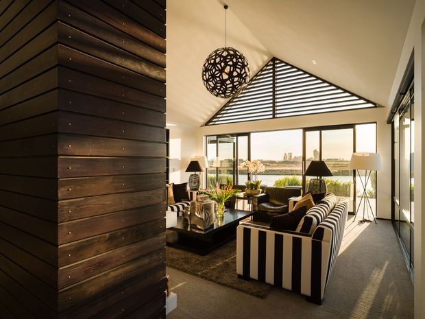 A sophisticated living room with expansive floor-to-ceiling glazing overlooking a waterfront or harbour setting. The room features a large striped sofa, dark lounge chairs, a low black coffee table, floor lamps, and a sculptural spherical pendant. A dramatic triangular clerestory window with horizontal louvres forms a strong architectural feature, while dark horizontal timber wall cladding contrasts with the warm neutral interior.