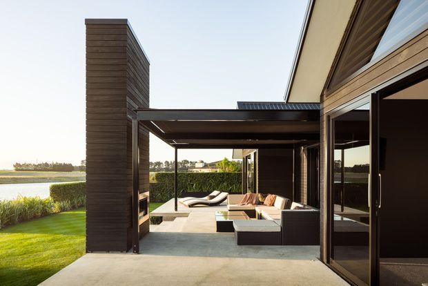 Exterior view of a contemporary dark-clad house with a covered outdoor entertaining area opening onto a manicured lawn and waterfront landscape. The pavilion-like terrace includes built-in seating, outdoor loungers, a low table, large sliding glass doors, and strong horizontal roof and wall lines.