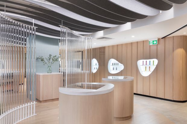 Contemporary commercial interior, likely a showroom or reception space, with curved light-wood display counters, integrated illuminated product niches, vertical metal rod screens, and a dramatic undulating dark ceiling feature. The space has pale timber wall panelling, light flooring, recessed lighting, and a clean, refined fit-out.