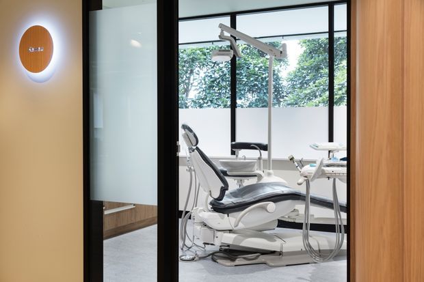 A bright, contemporary dental treatment room viewed through black-framed glass doors. The space contains a white dental chair, overhead examination light, dental equipment, and cabinetry, with large windows providing views of greenery. Warm wood wall panelling and cabinetry contrast with the cool grey flooring and white clinical fixtures.