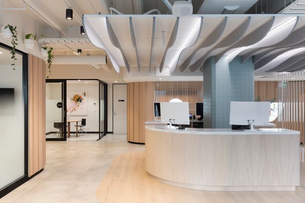 A bright contemporary commercial reception area with a large curved light-wood reception desk, white countertop, two computer workstations, glass-partitioned rooms, pale timber wall panelling, a teal tiled column, indoor plants, and a distinctive sculptural wave-like acoustic ceiling installation with integrated lighting. The space has an open, refined workplace or healthcare-office feel.