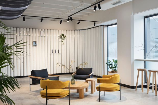 A bright contemporary commercial lounge or reception area with sculptural mustard-yellow lounge chairs, dark upholstered seating, and round timber coffee tables arranged on a pale tiled floor. The space features a ribbed vertical feature wall, extensive glazing, indoor plants, exposed white ceiling services, and black track lighting. A small high table with stools sits beside the windows.