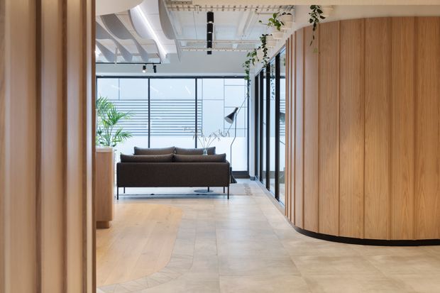 Contemporary office or commercial interior with a refined reception and lounge area. A dark sofa sits against full-height glazing, while a sweeping curved timber-clad wall and integrated cabinetry define the right side. The space features pale timber and stone flooring, exposed ceiling services, black-framed glazing, track lighting, and trailing greenery, creating a warm yet highly modern workplace environment.