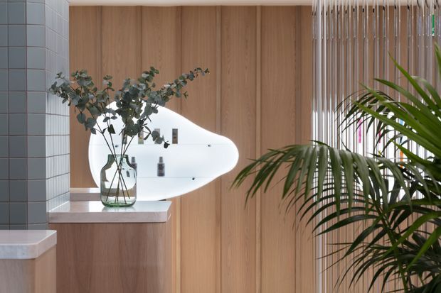 Contemporary bathroom interior featuring warm vertical timber wall panelling, pale stone vanity benchtops, a curved white integrated basin or wall feature, grey square wall tiles, and a slatted screen beside lush indoor planting. A vase of eucalyptus branches adds a natural accent.