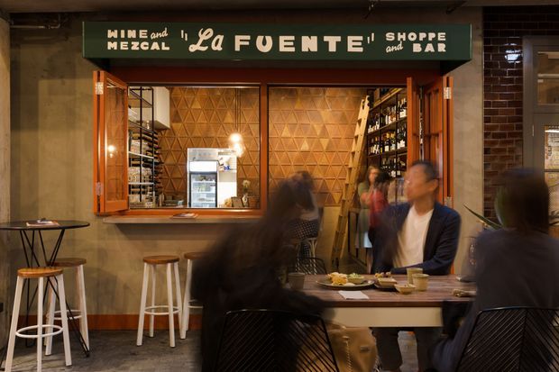 An intimate wine and mezcal bar with a service counter, open bottle shelving, geometric timber wall panelling, warm pendant lighting, and casual dining seating. The image includes patrons seated at tables and stools, with a dark green illustrated sign above the bar.