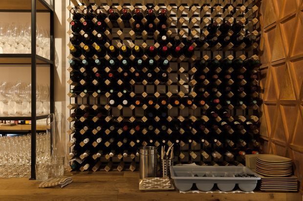 A richly detailed wine storage and serving area dominated by a large floor-to-ceiling geometric wine rack filled with bottles. Open black-framed shelving with glassware appears on the left, while a warm timber geometric panel forms the backdrop on the right. The timber countertop holds bar accessories, a metal container, serving items, and stacked plates. The overall setting is dark, warm, and sophisticated.