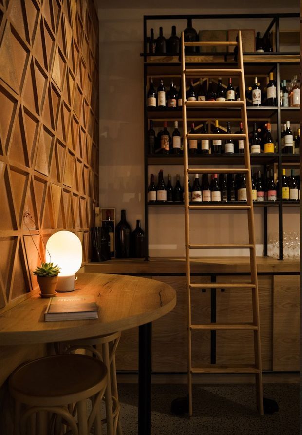 Warm, moody wine storage and tasting area with floor-to-ceiling dark shelving filled with wine bottles, a timber rolling ladder, wood cabinetry, a round timber table, stools, and a softly glowing lamp. The wall features a geometric timber screen, while terrazzo-like flooring and dark metal shelving supports add texture and contrast.