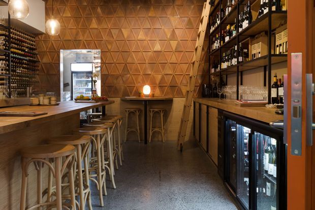 A narrow, warmly lit wine bar or hospitality space with a long timber counter and rows of stools on the left, extensive dark shelving stocked with wine bottles and glassware on the right, under-counter beverage refrigerators, and a striking geometric timber feature wall at the rear. Pendant lights and amber illumination create an intimate, richly textured atmosphere.