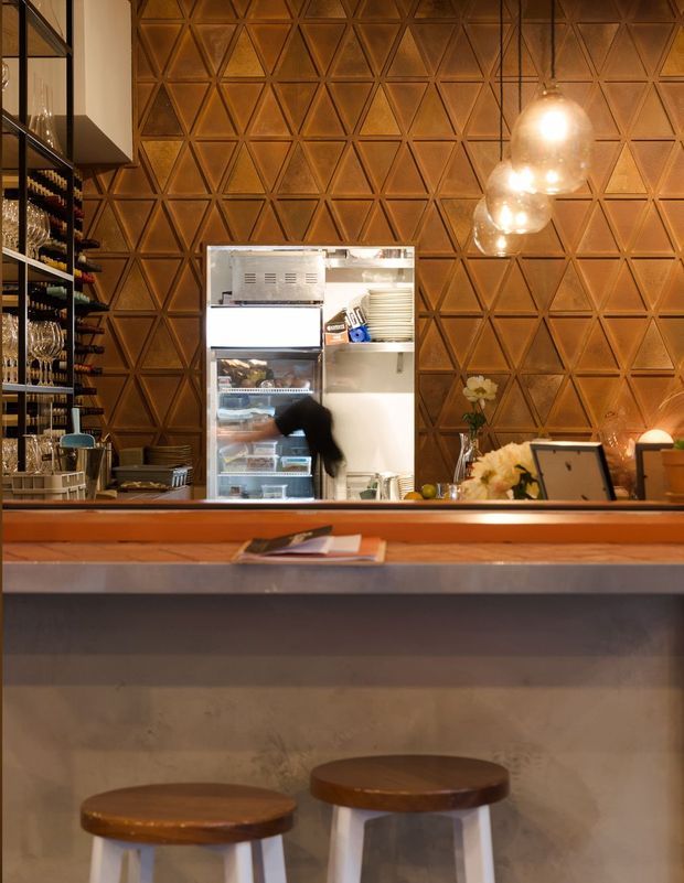 A warmly lit hospitality bar or café interior viewed from the customer side. The foreground features a concrete-faced counter with a dark metal edge and two wooden-topped stools. Behind the counter are open shelving stocked with glassware and bottles, a commercial refrigerator, pendant globe lights, and a striking geometric triangular timber-toned wall surface.
