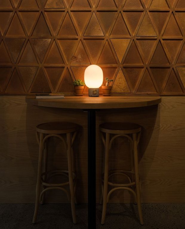 A dimly lit, intimate bar-height seating nook with a curved timber counter, two bentwood stools, and a glowing table lamp. The wall behind features a geometric triangular embossed panel in warm brown tones, with timber wall panelling below and small potted plants on the counter.