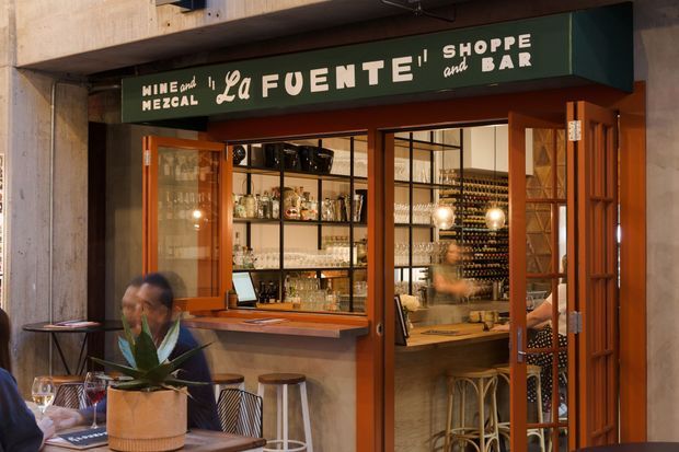 Street-facing entrance to La Fuente wine shop and bar, with warm timber-framed glazed doors and windows, a dark green sign, exposed concrete structure, and a visible interior featuring bottle displays, shelving, a bar counter, and stools.