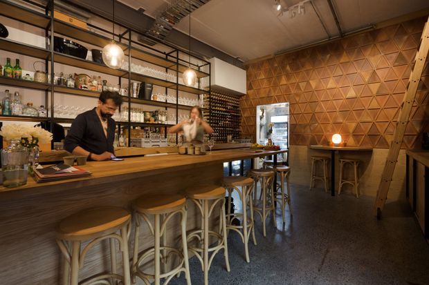 Warmly lit contemporary bar or hospitality interior with a long timber counter and stools, open metal-framed shelving displaying bottles and glassware, a wine rack, service area, and a striking geometric timber feature wall. The space has an industrial fit-out with exposed ceiling services, pendant lighting, polished concrete flooring, and a ladder leading to upper storage.