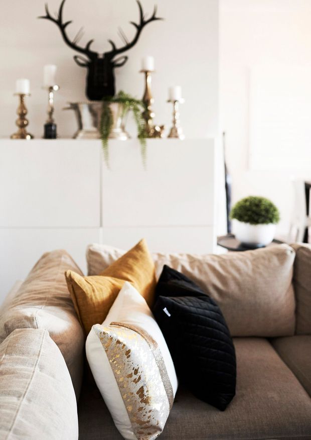 Bright, softly styled living room vignette featuring a large beige sectional sofa layered with cream, mustard-gold, and black cushions. A white built-in cabinet behind the sofa displays decorative candlesticks, a black antler sculpture, a metallic planter with trailing greenery, and other ornaments. A small round topiary is visible on a side surface, contributing to the refined neutral-and-gold scheme.
