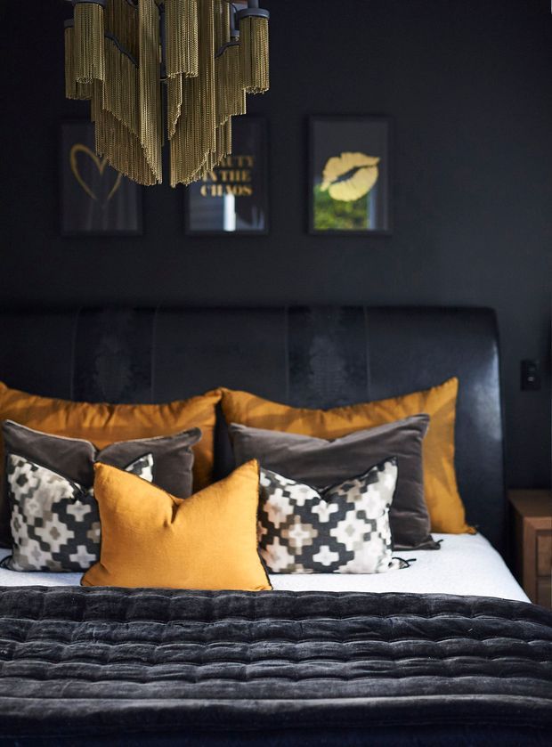A moody, luxurious bedroom featuring a dark upholstered bed with a tall channel-tufted headboard, layered black, charcoal, white, and mustard-gold cushions, and a plush dark quilt. A dramatic gold-toned chain pendant light hangs overhead, with framed artwork on the charcoal wall and a small bedside table at right.