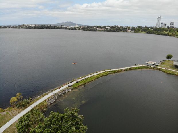 Aerial view of a broad waterfront landscape with a curving paved promenade and landscaped shoreline in the foreground. Calm water dominates the scene, with residential development, high-rise buildings, and a low hill visible across the water under a partly cloudy sky.