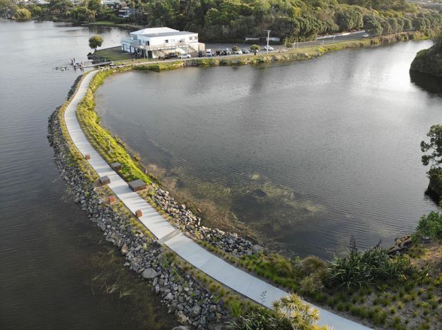 Aerial view of a landscaped waterfront site with a curving concrete pedestrian path along a rocky shoreline and narrow causeway. Calm water surrounds the path, with planted native-looking vegetation, a low white waterside building, roads, and dense greenery in the background.