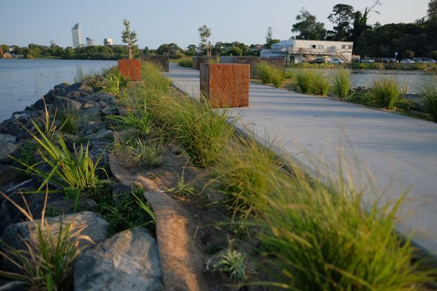 A landscaped waterfront promenade extends along a rocky shoreline, with a broad smooth concrete path, rust-coloured perforated metal screens or sculptural elements, and native grasses and planting. Calm water, low buildings, and a distant urban skyline are visible beyond the path.