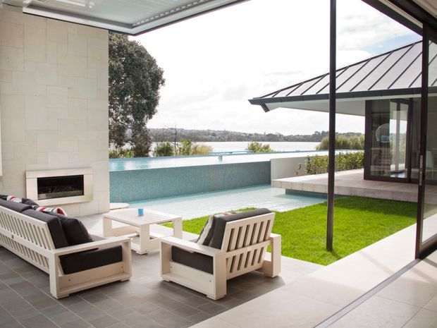 A refined covered outdoor living area opening onto an infinity-edge swimming pool and landscaped lawn, with a built-in fireplace, outdoor lounge furniture, large-format tiled flooring, and expansive glazing. The view overlooks a body of water and wooded landscape. A separate pavilion with a standing-seam metal roof is visible across the courtyard-like pool area.