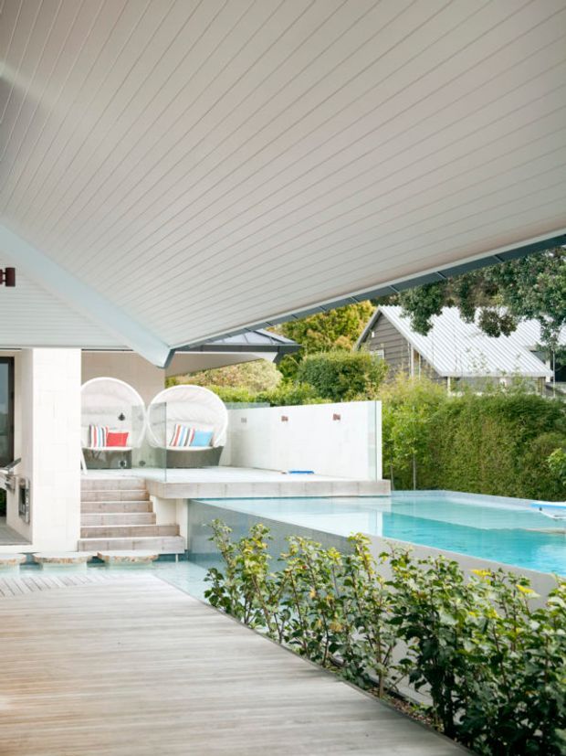 A bright, contemporary outdoor entertaining area viewed from beneath a broad white-lined roof canopy. The space features a timber deck, a turquoise swimming pool with integrated shallow platform, pale stone or tiled steps, built-in seating, and sculptural round lounge chairs. A white privacy wall and landscaped hedging frame the pool, with neighbouring pitched-roof structures visible beyond.