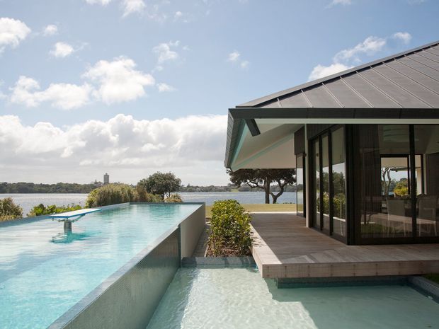 Contemporary waterfront residence with a low-pitched metal roof, extensive dark-framed glazing, and a timber deck beside an infinity-edge swimming pool. A shallow pool or water feature extends around the deck, with a calm body of water and distant shoreline visible beyond the property.
