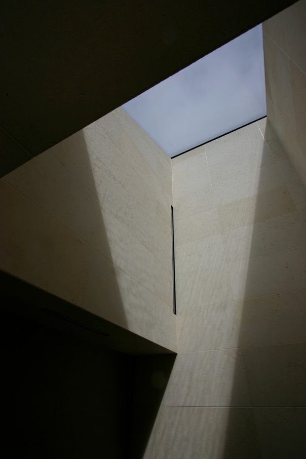 Dramatic upward view of a tall, narrow interior void with pale stone or concrete wall panels and a large overhead skylight. Strong natural light creates angular shadows across the surfaces, emphasizing the minimalist geometric architecture.