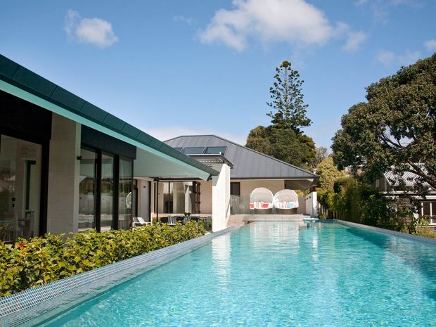 Exterior view of a contemporary residential property centered on a large rectangular swimming pool. The home has low, angular rooflines, dark metal roofing, extensive black-framed glazing, pale rendered walls, and a covered outdoor entertaining area. Landscaped planting borders the pool, with mature trees surrounding the property.