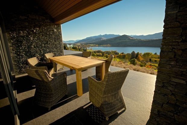 Covered outdoor dining terrace with a timber dining table and woven wicker chairs, framed by dark stacked-stone walls and a warm timber-lined ceiling. The open view looks across a lake toward surrounding mountain ranges, with a broad paved floor and landscaped foreground.