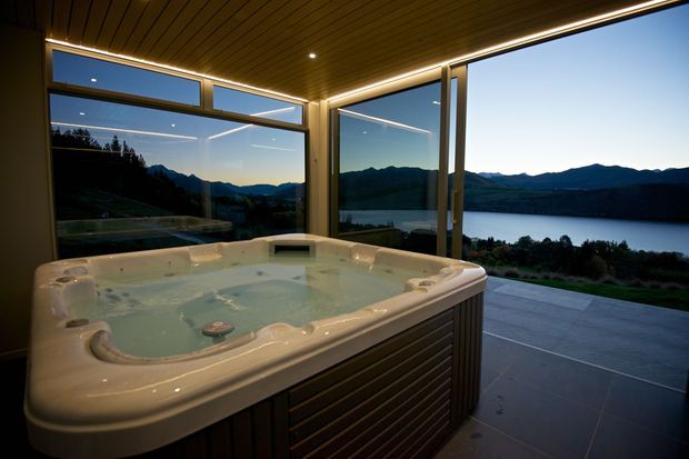 A luxurious indoor spa room with a large freestanding white hot tub positioned beside expansive floor-to-ceiling glazing. The room has a dark timber slatted ceiling with integrated linear lighting, dark-framed glass walls and doors, and a tiled floor. Through the glazing is a dramatic mountain and lakeside landscape at dusk.