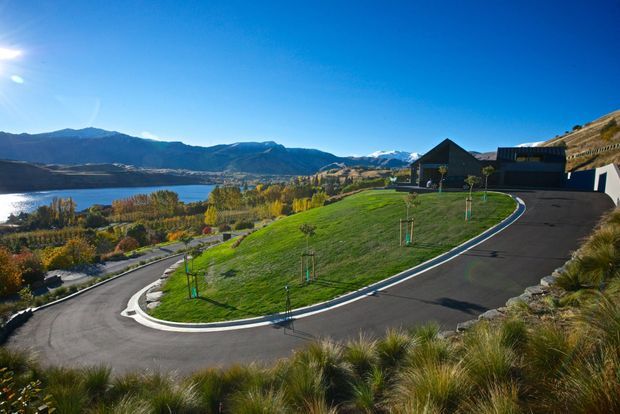 A wide exterior view of a contemporary dark-clad house set on a landscaped hillside above a lake, with a sweeping paved driveway, newly planted trees, ornamental grasses, and mountain scenery in the background. The property appears to occupy a sloped, elevated site with a pitched-roof main volume and connected flat or low-pitched sections.