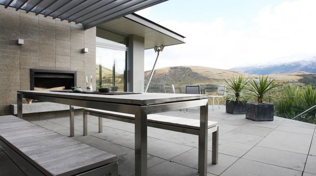 Contemporary outdoor entertaining terrace with a large stainless-steel dining table and bench seating beneath a slatted pergola. The terrace features pale large-format paving, a built-in outdoor fireplace set into a textured stone wall, minimalist roof and glazing, and views toward a rugged mountainous landscape.
