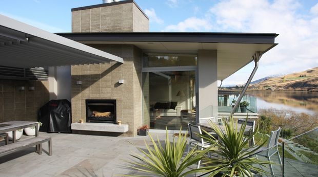 Contemporary lakeside home with a broad flat roof and cantilevered outdoor terrace. The image focuses on a paved outdoor entertaining area featuring a built-in fireplace, dining table, barbecue, outdoor seating and sculptural planted greenery. Floor-to-ceiling glazing reveals a bright interior living area, while the calm lake and surrounding hills form the background.