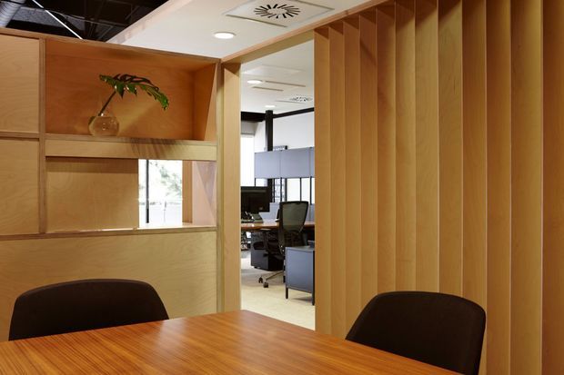 A warm timber-lined meeting or conference room with a large wood table and dark upholstered chairs. Built-in timber shelving and a vertically slatted timber partition frame an opening to a modern office workspace beyond. Recessed ceiling lighting and exposed ceiling services are visible.