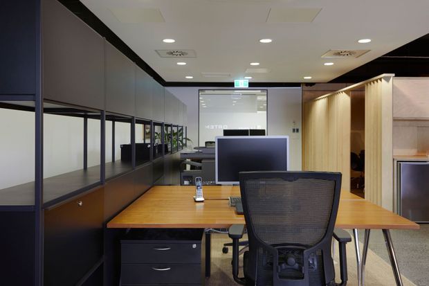 Contemporary open-plan office interior with shared timber workstations, black mesh office chairs, dark-framed glazed partitions, integrated storage, recessed ceiling lighting, and a timber slatted meeting or kitchenette zone at the rear.