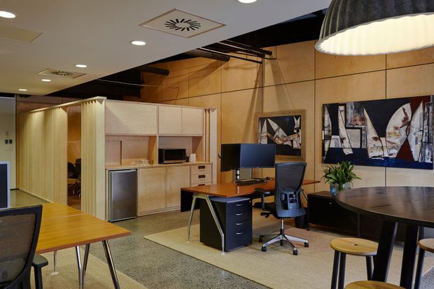 A warm, open-plan office interior with timber wall panelling, built-in light-wood kitchenette and storage cabinetry, individual workstations, a meeting table, and contemporary pendant and recessed lighting. Abstract artwork and office furniture create a refined mid-century-influenced commercial workspace.