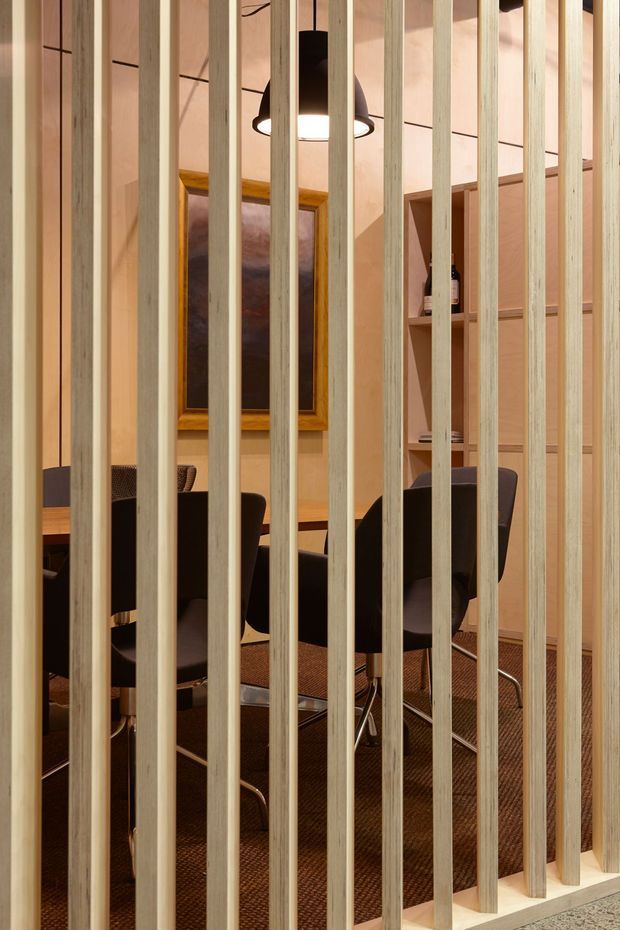 A warm, contemporary dining or meeting area viewed through a vertical timber slat screen. The space contains a dark table and several upholstered chairs, a framed artwork, open timber shelving, pendant lighting, and a textured brown carpet. Pale timber battens create a semi-transparent partition across the foreground.