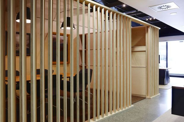 Contemporary commercial interior featuring a semi-enclosed meeting or dining area screened by closely spaced vertical timber battens. The space includes dark upholstered chairs, a timber table, pendant lighting, built-in timber joinery, polished concrete or terrazzo-style flooring, and an exposed dark ceiling with integrated services. Bright glazing is visible in the background.