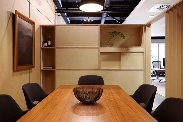 A contemporary office meeting room with a large rectangular timber table, dark upholstered chairs, and a full-height timber built-in storage and display wall. The space features warm wood finishes, a framed artwork, exposed black ceiling services, recessed lighting, and glazing opening to an adjacent office area.