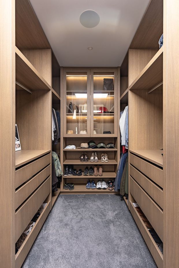 A compact walk-in wardrobe lined with warm wood-grain built-in cabinetry on both sides. Open shelving, drawers, hanging rails, and a central glass-front display cabinet provide organized clothing, accessories, and extensive shoe storage. The room has a grey carpeted floor, a smooth white ceiling with recessed lighting, and integrated warm LED illumination in the shelving.