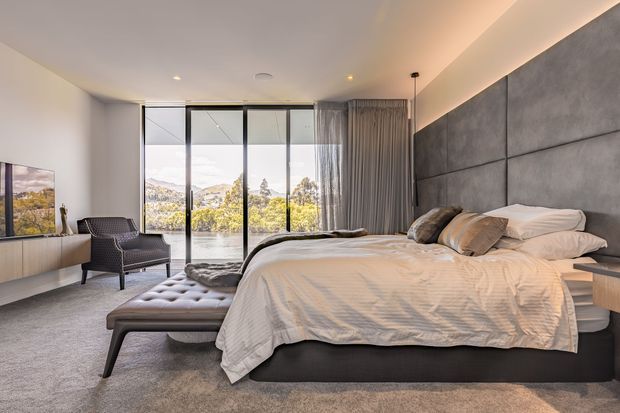 A spacious contemporary bedroom with a large upholstered bed, white bedding, a tufted bench, and a dark woven lounge chair. Floor-to-ceiling sliding glass doors open to a leafy suburban or hillside view, while sheer curtains provide privacy. The room features a dark grey padded feature wall, neutral carpet, pale built-in cabinetry, recessed ceiling lighting, and warm concealed cove lighting.