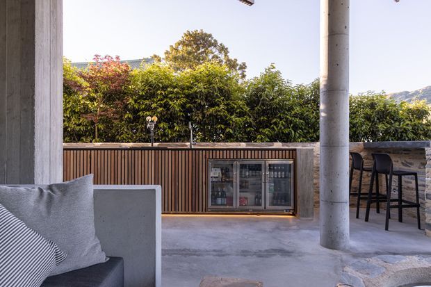 An outdoor entertaining area with a partially covered concrete terrace, built-in outdoor bar and beverage refrigerator, vertical timber screening, stone-clad counter, and high bar stools. A concrete column and outdoor lounge seating frame the space, with dense greenery and distant hills beyond.