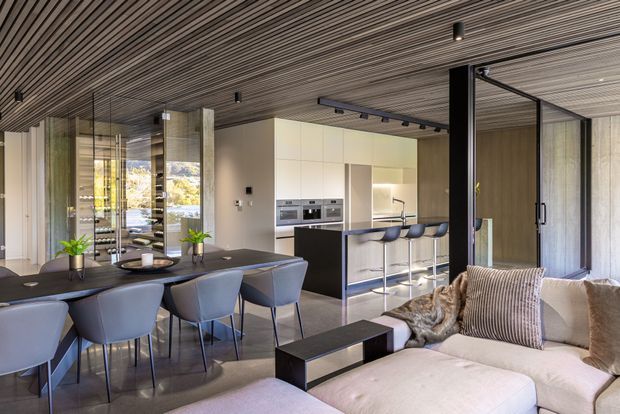 Open-plan contemporary interior combining a dining area, kitchen and living room. A large dining table with upholstered chairs sits in the foreground, facing a streamlined kitchen with a dark island, integrated appliances and bar seating. The living area includes a pale sectional sofa and low coffee table. Floor-to-ceiling glazing and a glazed partition bring in natural light, while the ceiling is clad in continuous narrow timber slats with recessed and track lighting. The palette is neutral, warm and refined, with dark cabinetry and framing providing contrast.