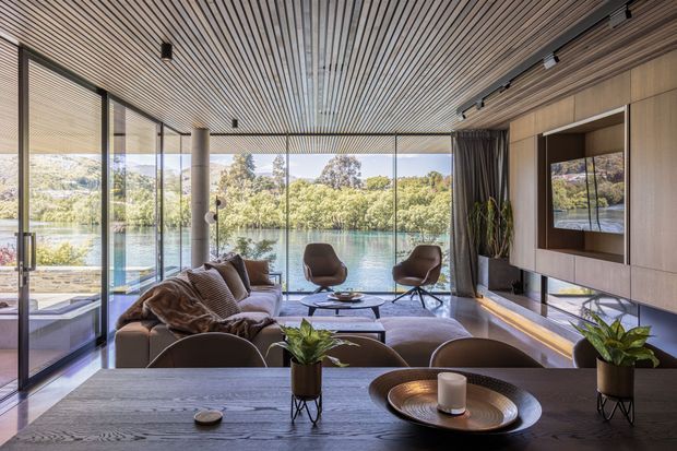 A refined contemporary living room with expansive floor-to-ceiling glazing overlooking a tranquil body of water and landscaped greenery. The room features a warm timber slatted ceiling, timber wall panelling with an integrated television, dark-framed glass doors, a neutral sectional sofa, sculptural lounge chairs, a low coffee table, and a dark timber dining table in the foreground. Natural light, layered textures, and restrained furnishings create a calm, luxurious indoor-outdoor atmosphere.