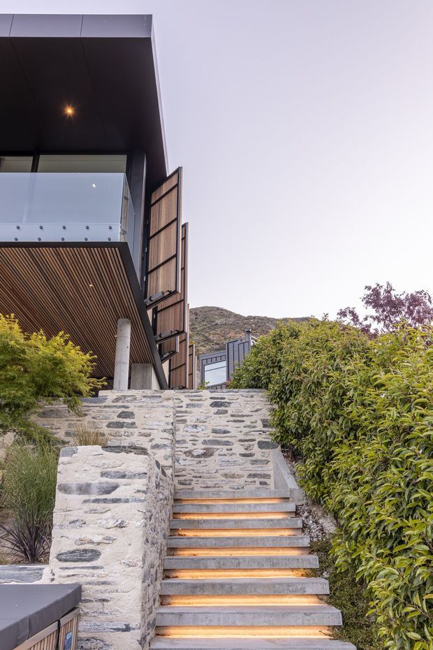 Exterior view of a contemporary multi-level building approached by a landscaped staircase. The structure features dark cladding, a deep overhanging roof, timber-lined soffits, a glass balustrade, and vertical timber screening. Rough stone retaining walls frame the illuminated concrete steps, with dense planting and a rocky hillside visible beyond.