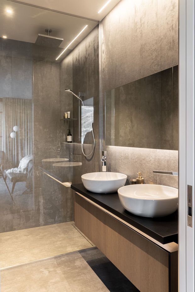 Contemporary bathroom with a walk-in shower, double vessel basins, wall-to-wall mirror, illuminated vanity, and large-format grey-brown stone or concrete-look surfaces. A glass partition reflects an adjacent bedroom area, while recessed linear lighting creates a refined, softly lit atmosphere.