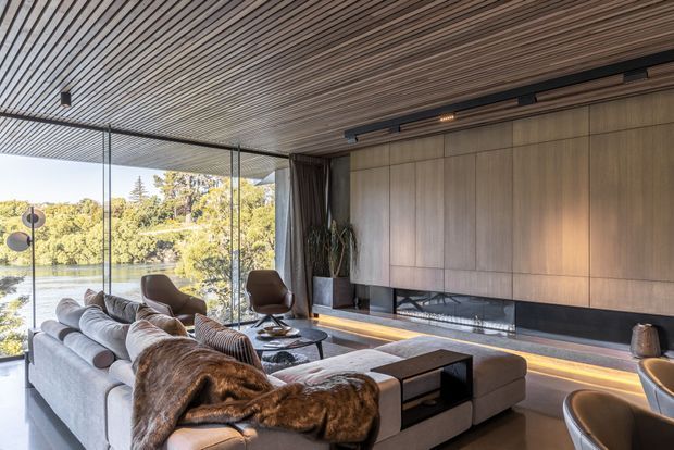 A sophisticated contemporary living room with a large sectional sofa, lounge chairs, low coffee tables, and a full-width built-in media wall. Floor-to-ceiling glazing opens the room to a leafy waterfront view, while warm linear lighting highlights the dark fireplace and timber-toned wall panelling. The ceiling features closely spaced linear timber battens, creating a refined and warm interior.