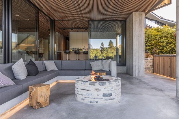 A contemporary covered outdoor lounge adjoining a modern home, with a large grey sectional sofa, round stone-clad fire pit, timber stool, concrete floor, exposed timber slatted ceiling, and extensive dark-framed glazing. An open kitchen is visible through the glass, while a landscaped hedge and timber gate appear at the side.