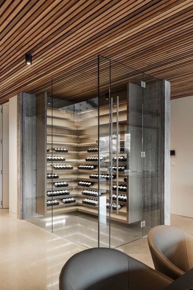A refined modern wine room enclosed by frameless glass walls and doors, with extensive built-in timber bottle shelving and concealed display lighting. The room features a linear timber slat ceiling, polished light flooring, dark concrete-look structural piers, and rounded leather seating in the foreground.