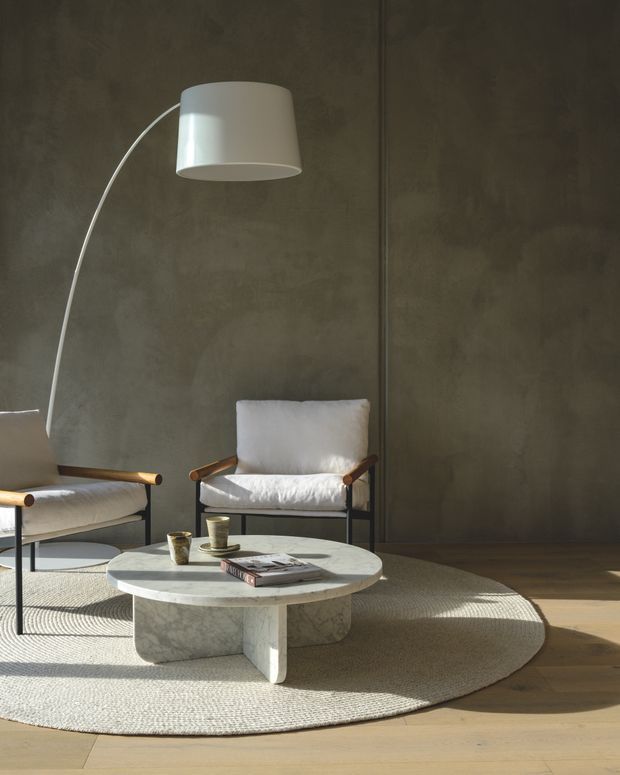 A softly lit contemporary living room with a dark textured plaster wall, pale timber flooring, two low lounge chairs with light upholstery and timber armrests, and a sculptural round stone coffee table. A large arched floor lamp, woven rug, coffee cups, and books complete the calm, minimalist seating area.